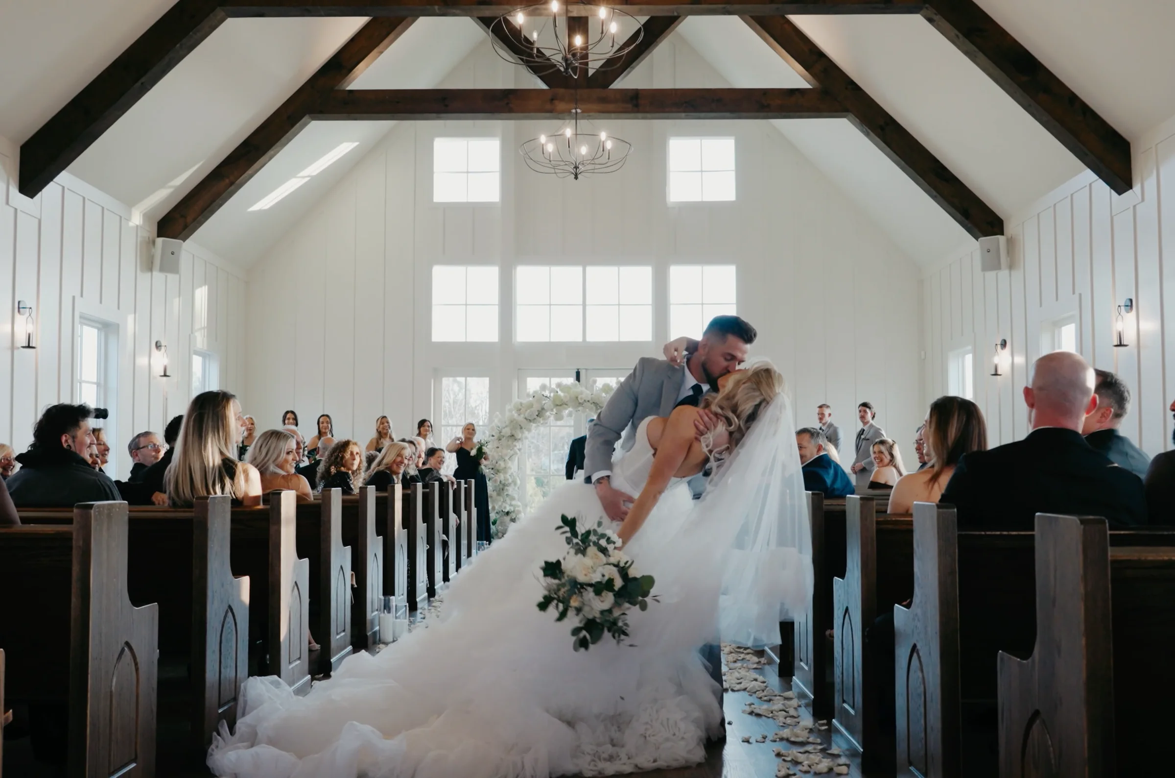 A groom dips a bride for a kiss in the aisle of a white chapel.
