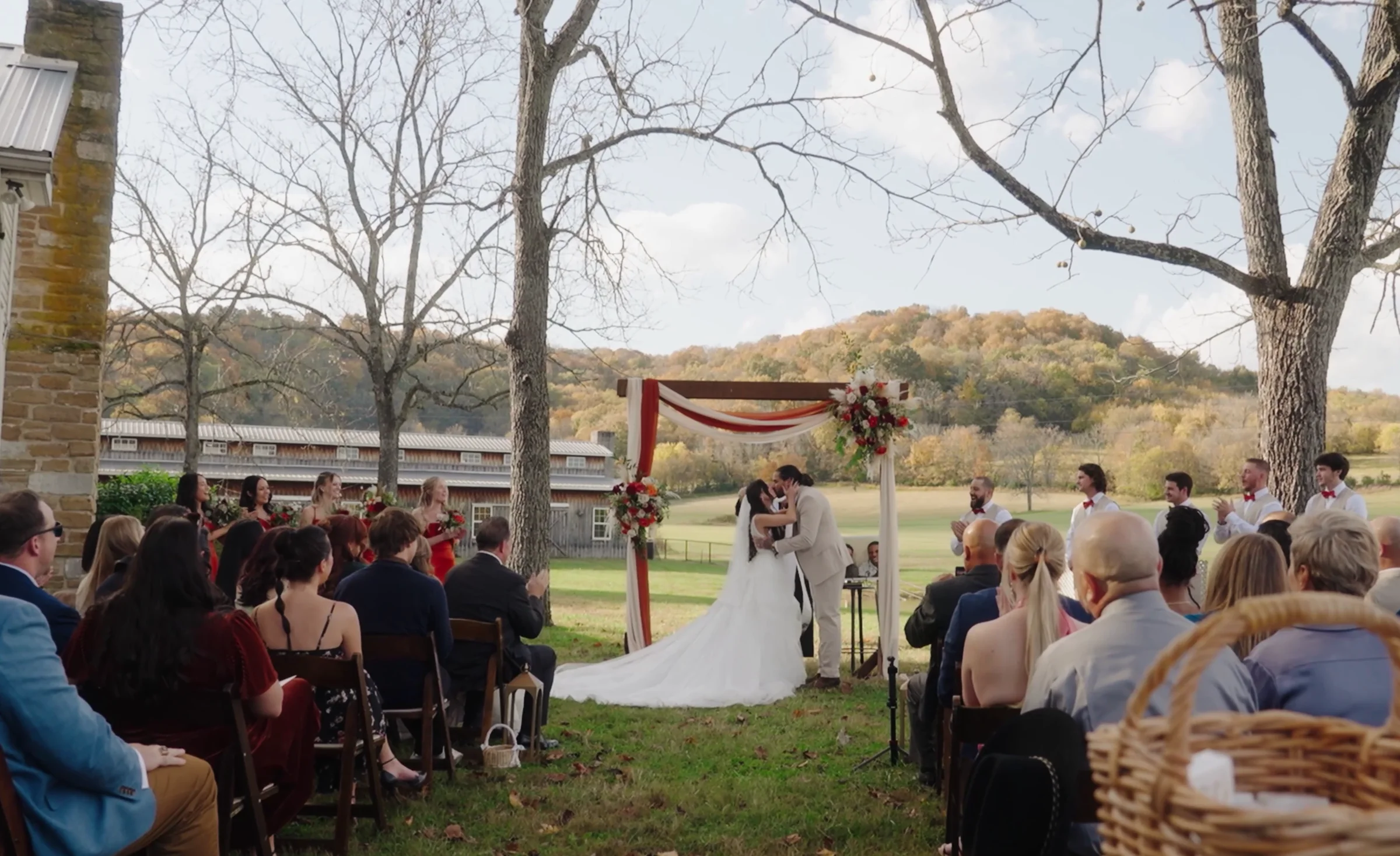 A couple kisses under a floral arch at an outdoor fall ceremony, guests seated on both sides