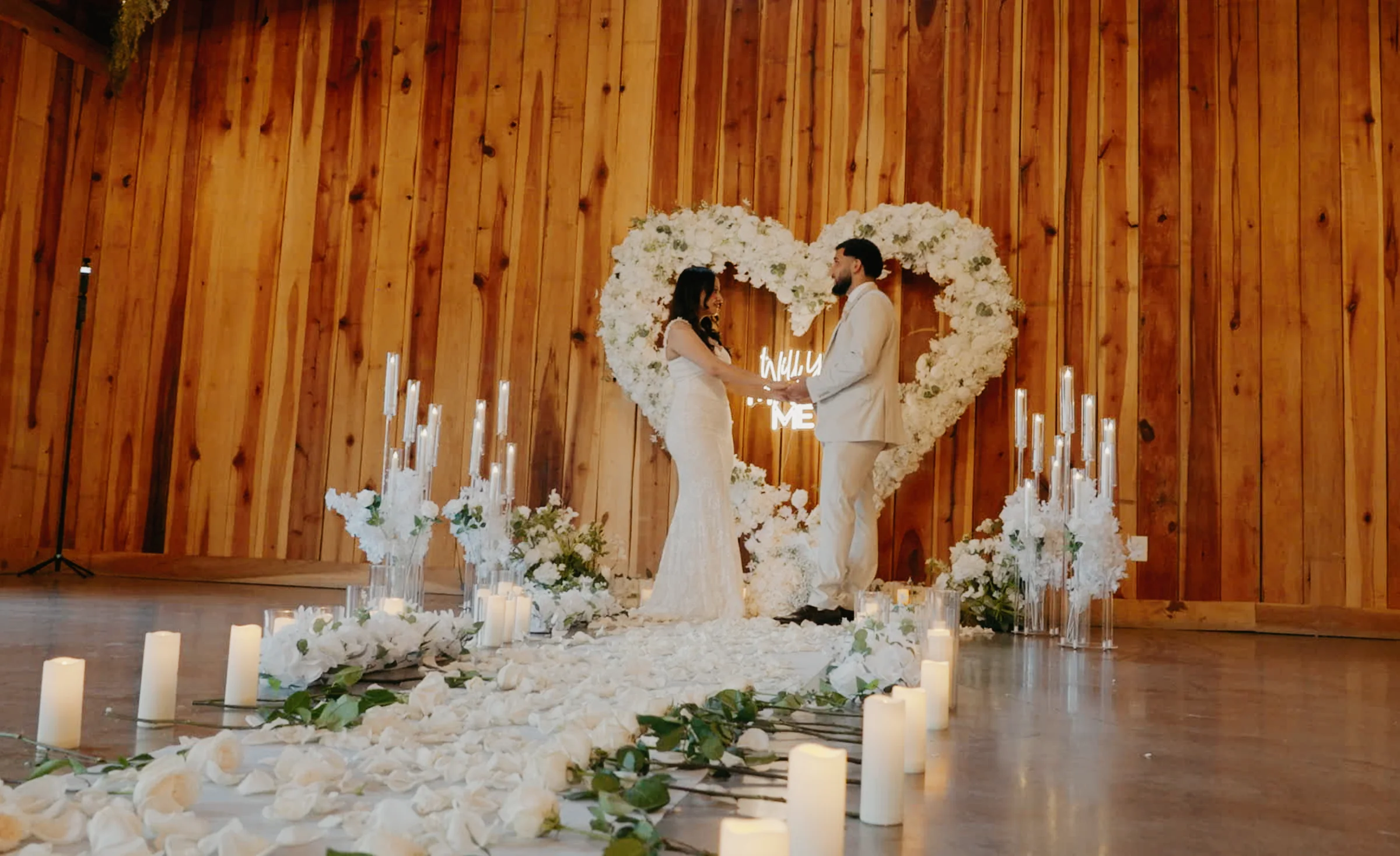 A couple stand alone under a heart-shaped floral arch inside a warm-lit barn.