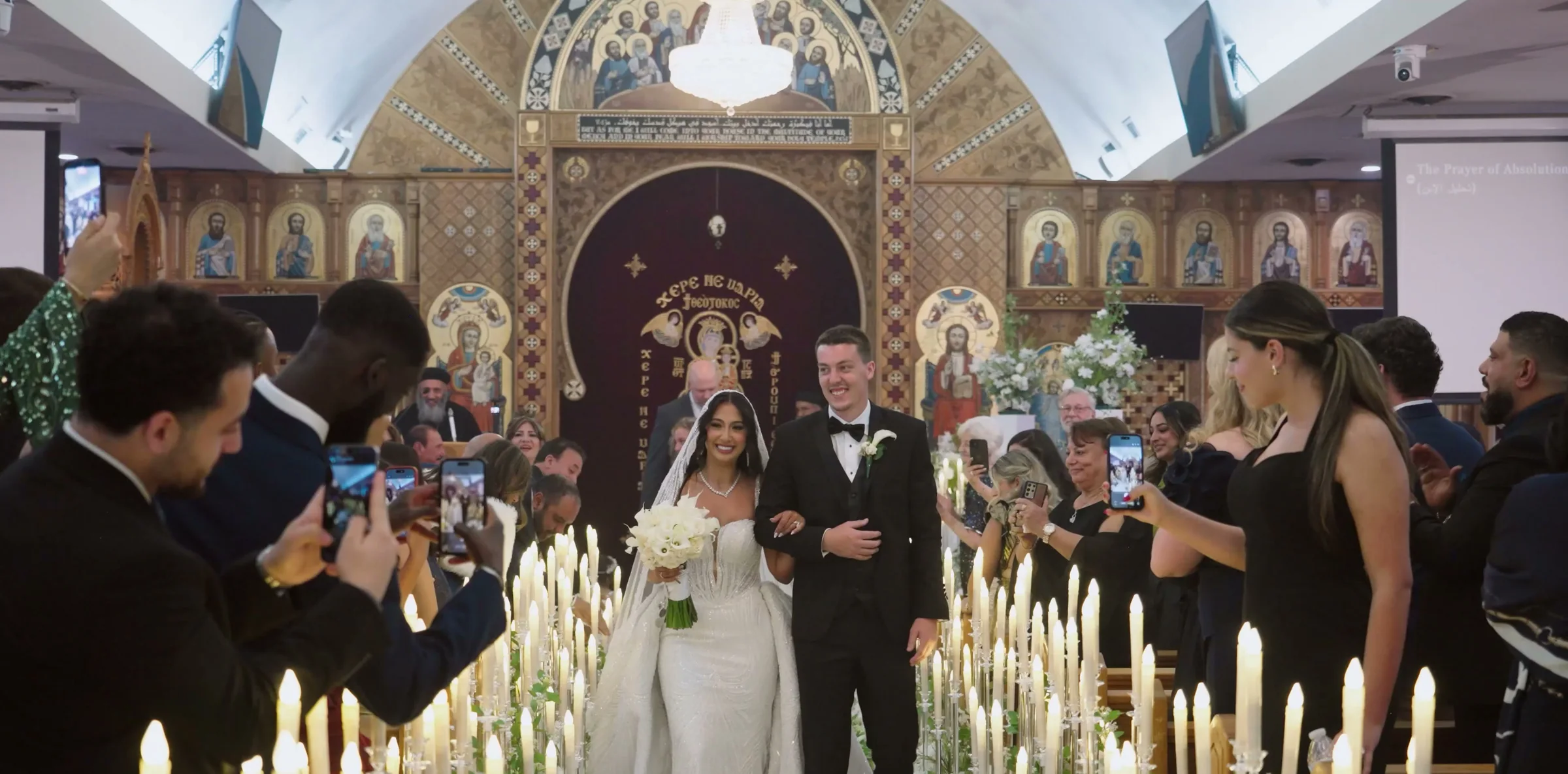 A couple walk back down a candle-lined aisle as guests film them.