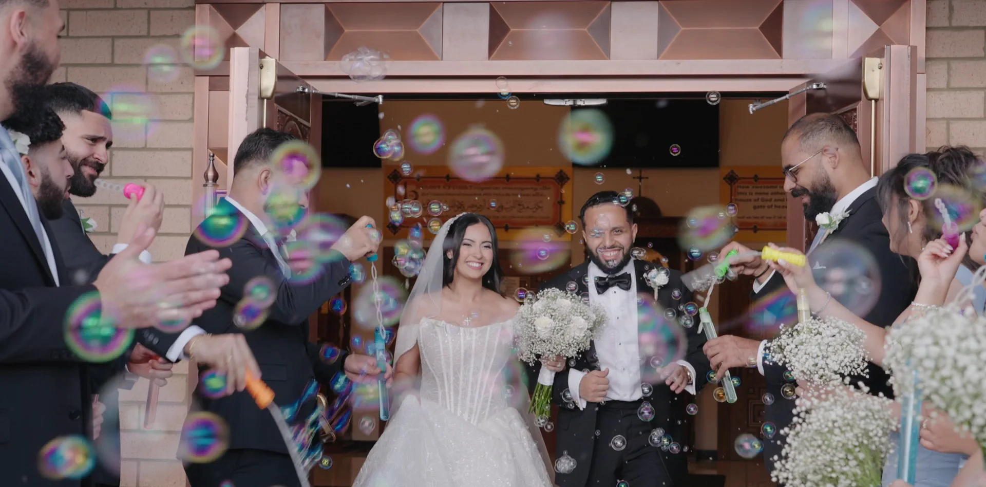 A couple walk out of a church doorway through a cloud of bubbles.