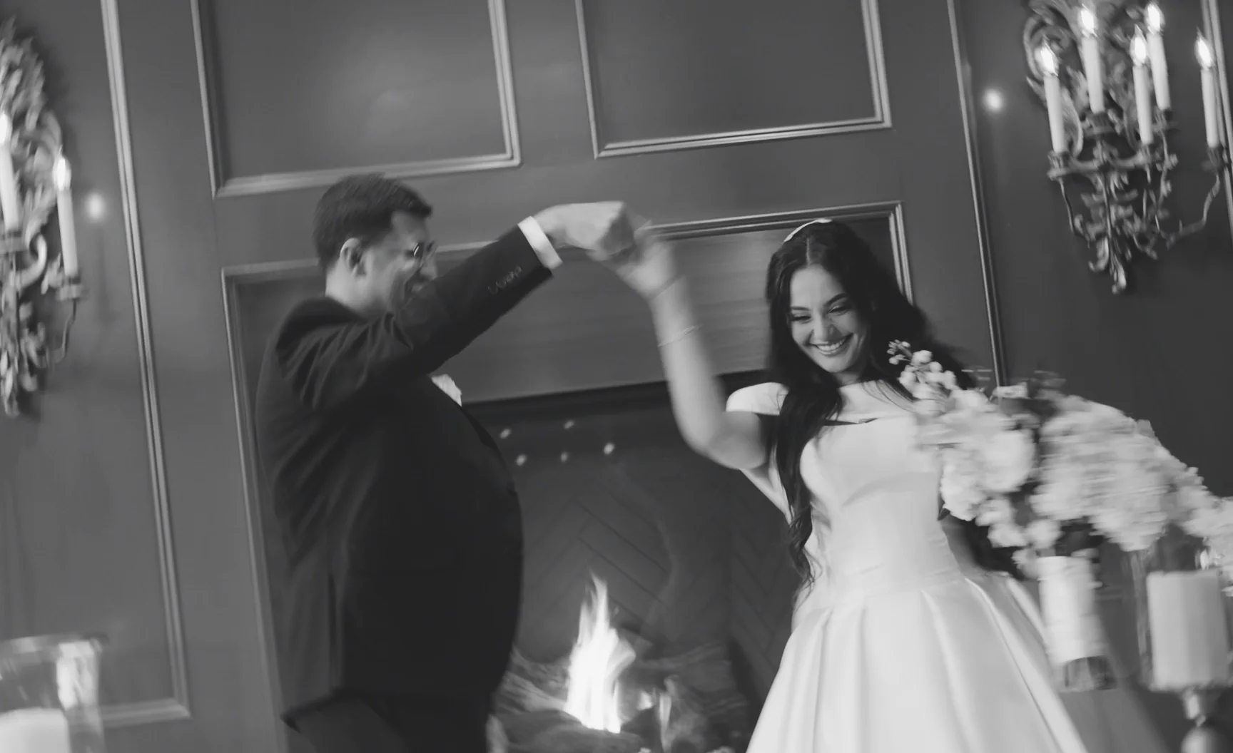 A groom spins a bride in front of a lit fireplace.