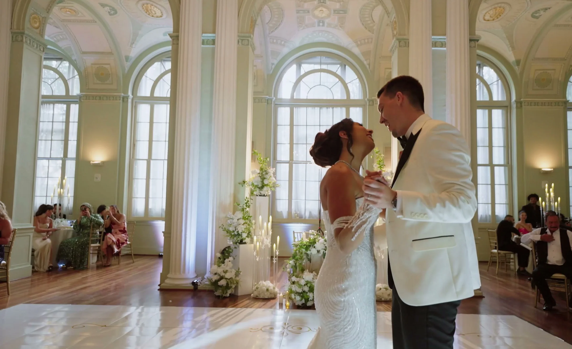 A couple dance in a grand hall with tall arched windows while guests watch
