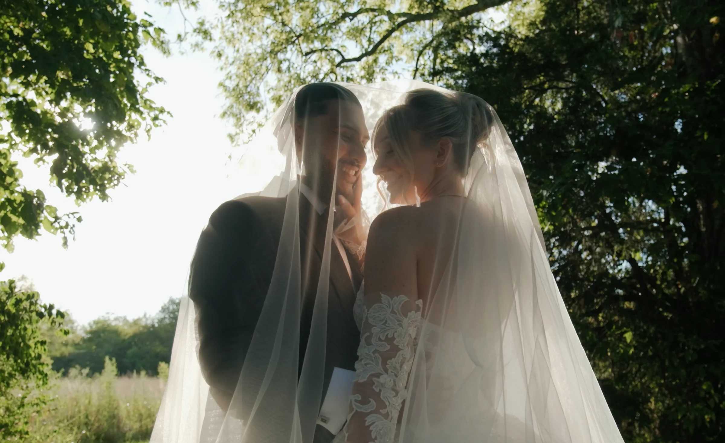 A bride and groom smile at each other under her veil, backlit by the sun through the trees