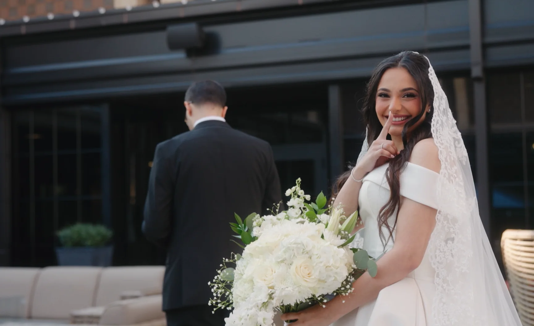 A bride raises a finger to her lips behind a groom who is facing away.