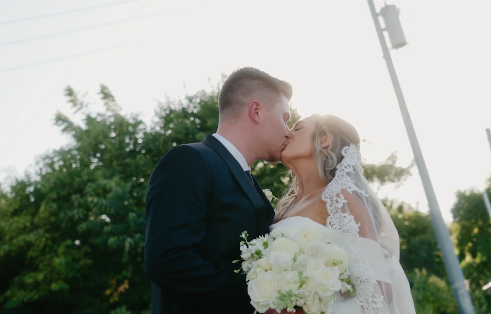 A couple kiss in golden evening light with a bouquet between them.