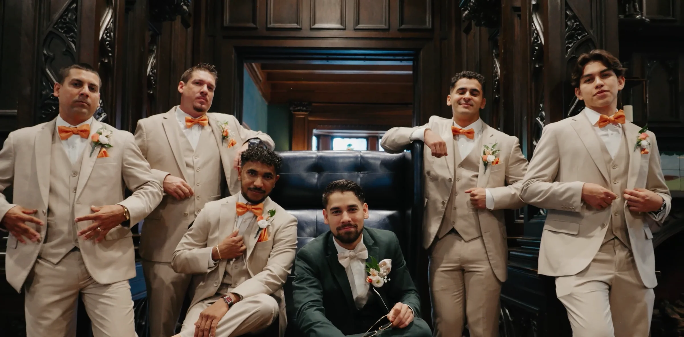 Groomsmen in tan suits pose around a seated groom in a wood-panelled room.