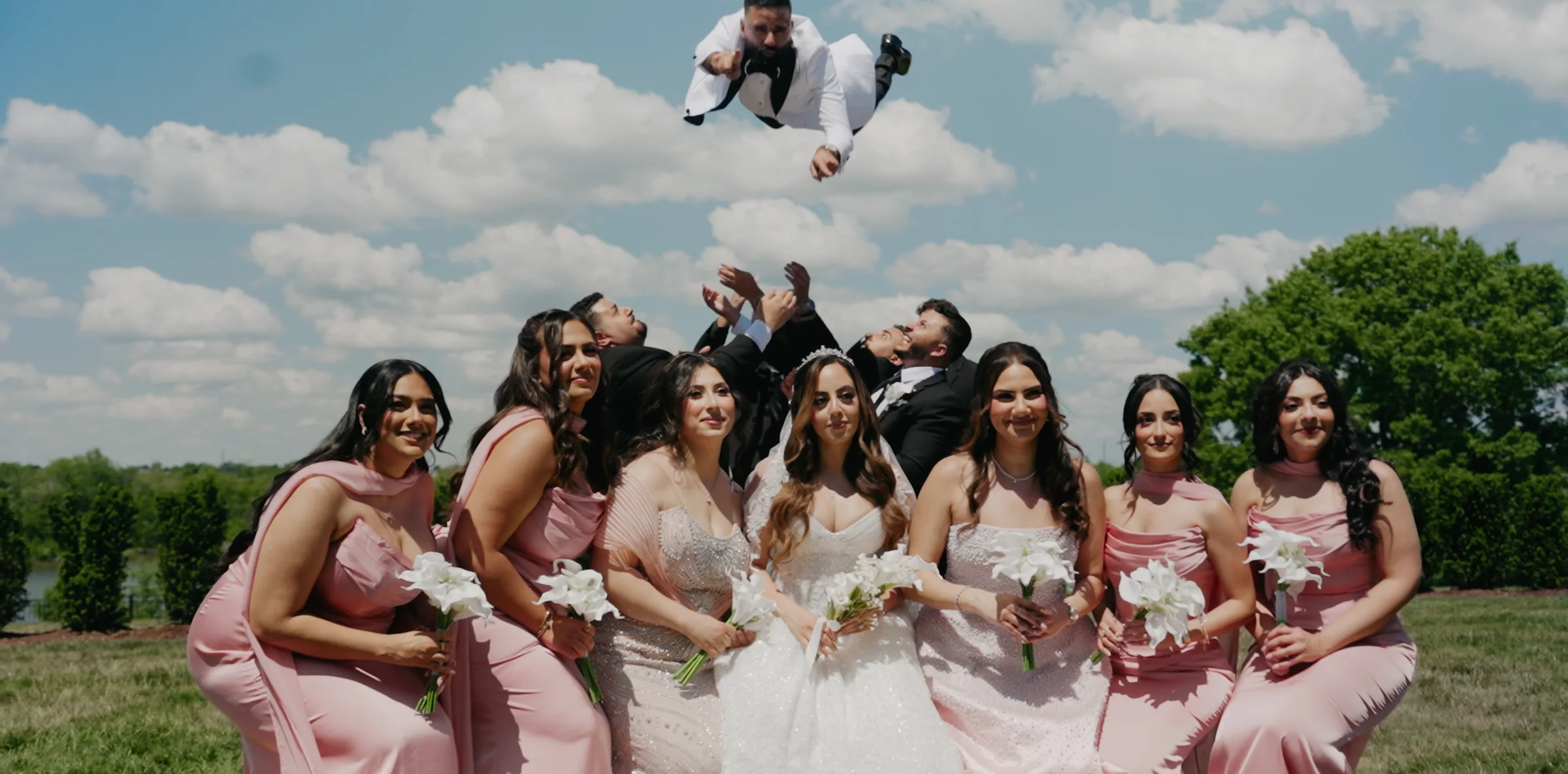A groom is thrown into the air above the bridal party against a blue sky.
