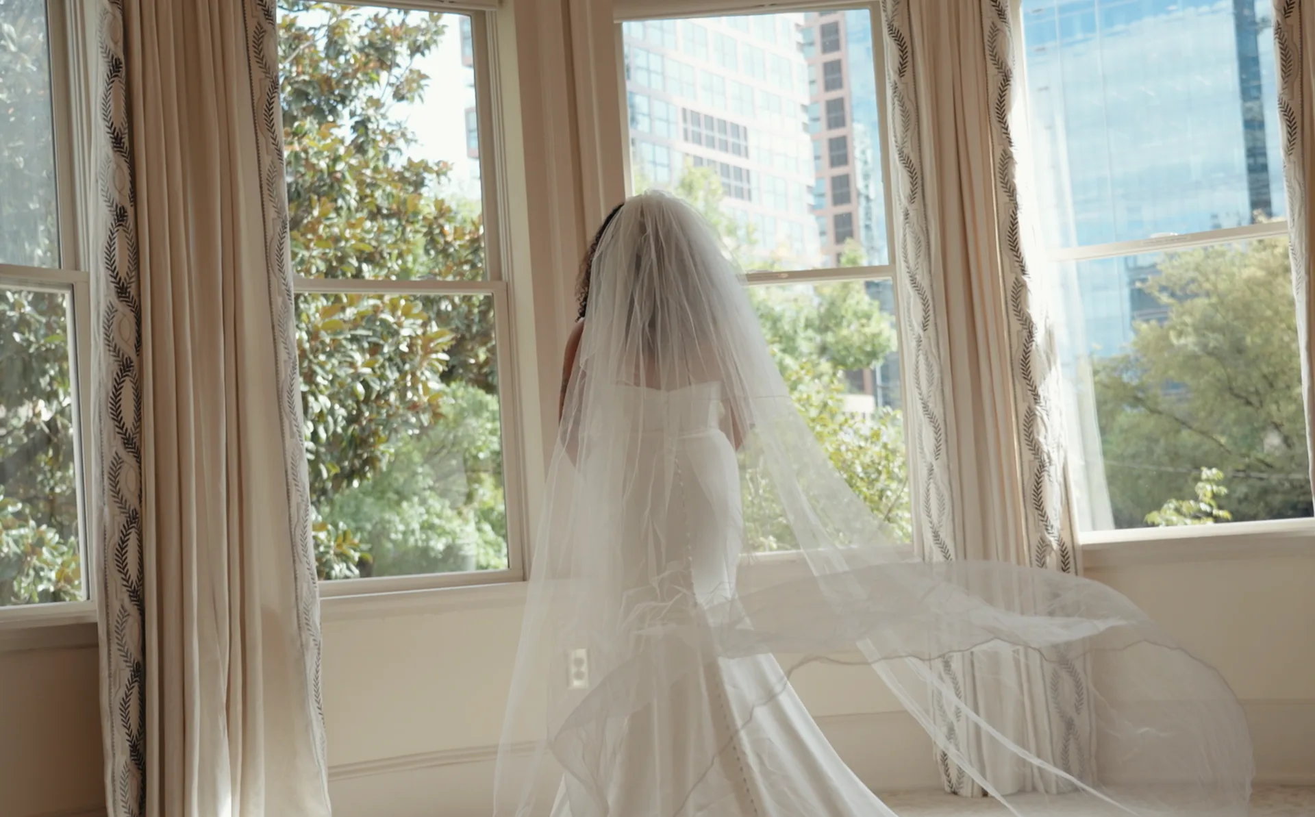 A bride stands at a bright window with her veil falling down her back.