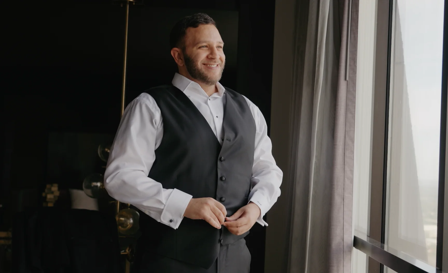 A groom fastens his vest by a window before the ceremony.