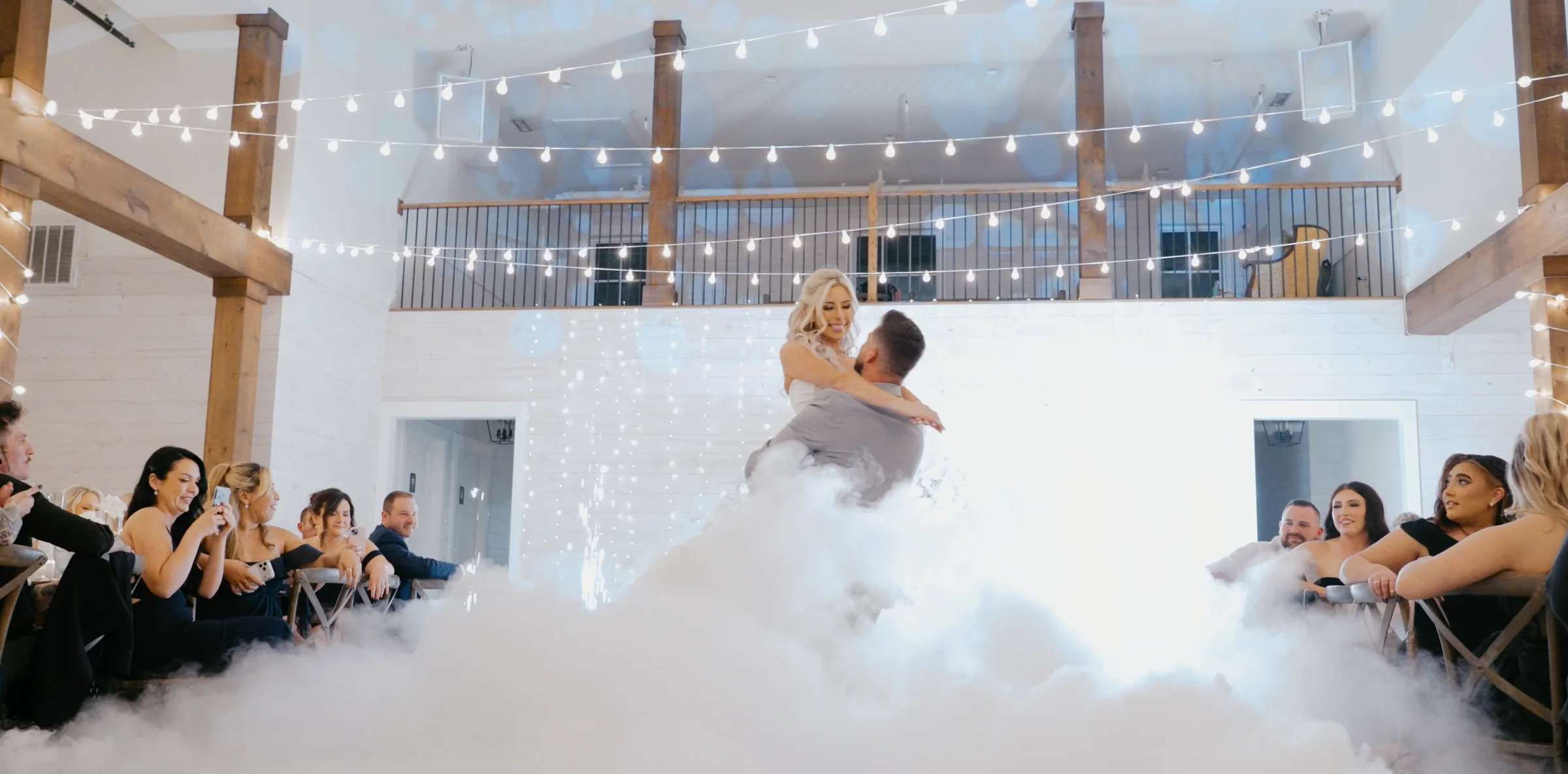 A groom lifts a bride above a low cloud of dry ice while guests watch.