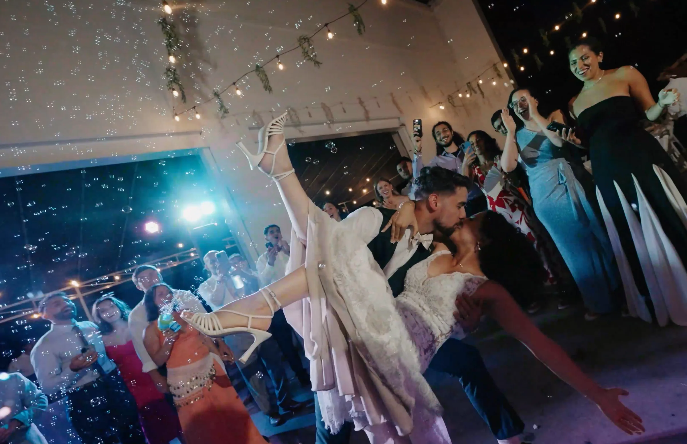A groom dips a bride for a kiss as bubbles fall and guests film.