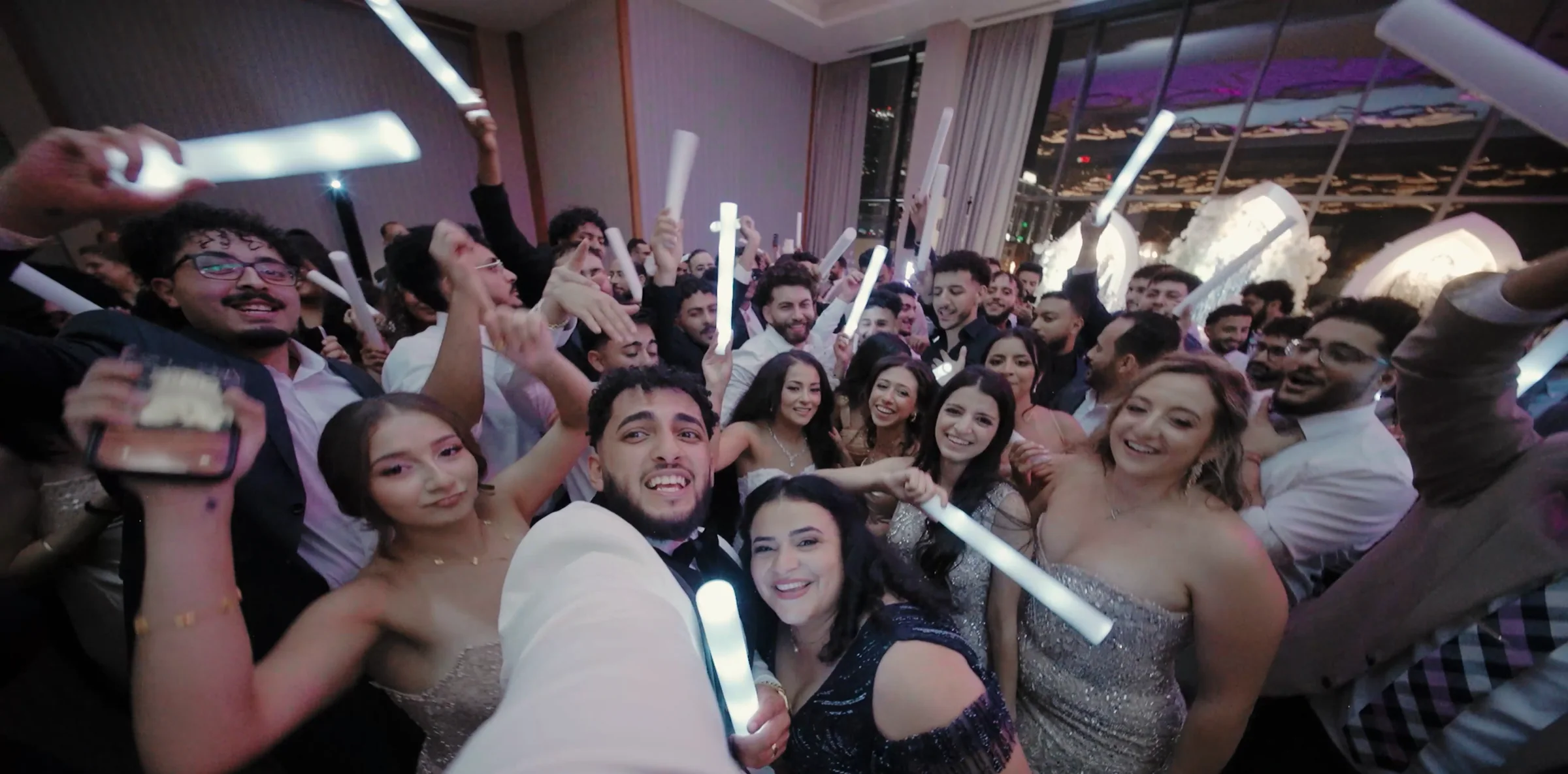 A groom holds the camera out for a selfie in the middle of a packed dance floor.