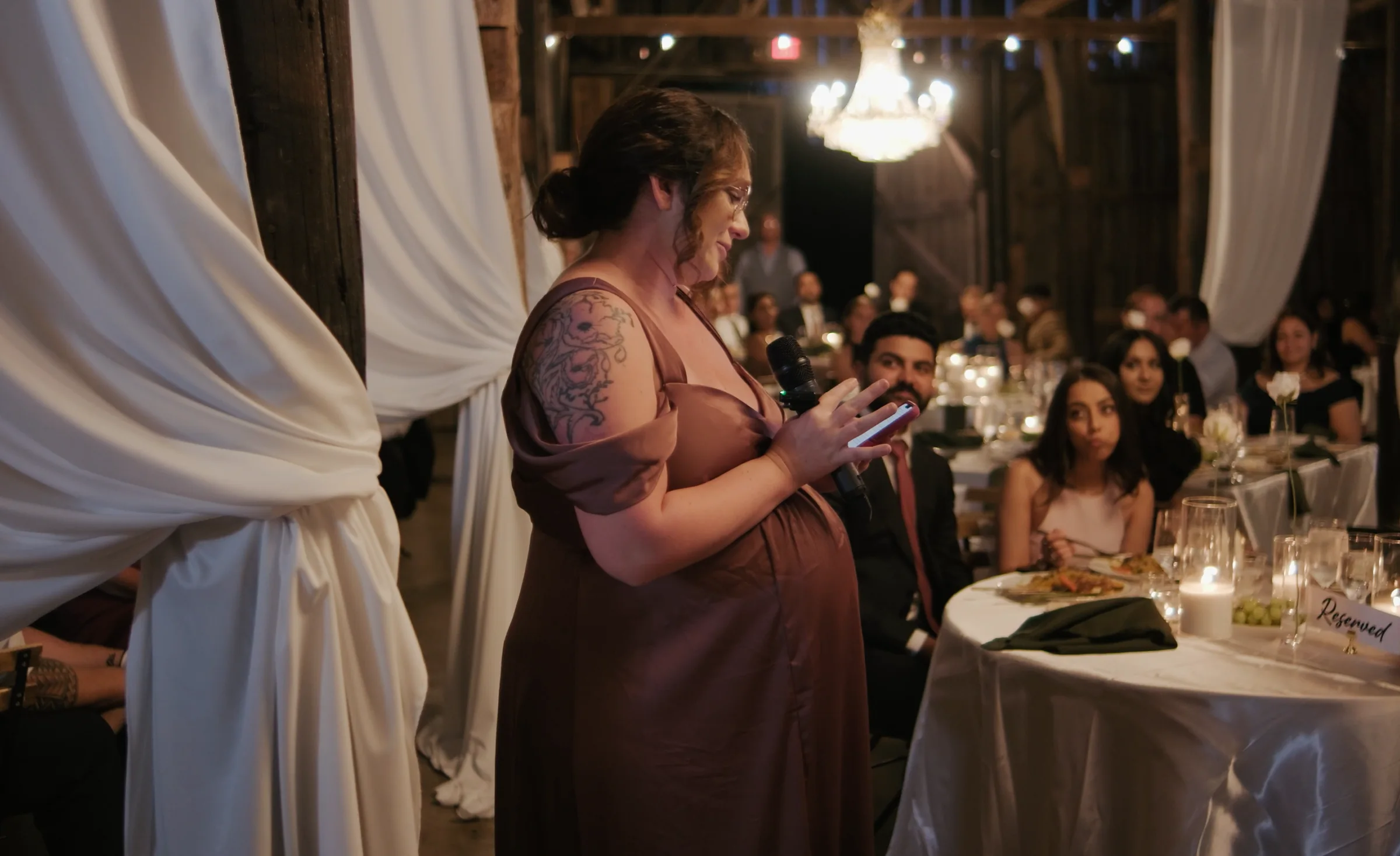 A woman reads her toast into a microphone at a candlelit reception while guests listen